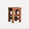 El Taburete bajo en madera de caoba envejecida Akviran presenta un asiento cuadrado con patas resistentes, elaborado en caoba y que muestra un acabado envejecido con un diseño en forma de bloques.