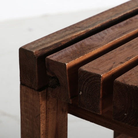 Primer plano de un taburete bajo de esquina Akviran hecho de madera de caoba envejecida, que muestra su diseño de listones y veta natural.