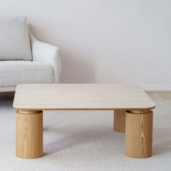 La Mesa de centro cuadrada en cerámico DM y fresno natural Línea realza un salón contemporáneo, elegantemente colocada sobre una alfombra beige y combinada con un sofá gris claro contra una pared lisa, mostrando la exquisita decoración del hogar.