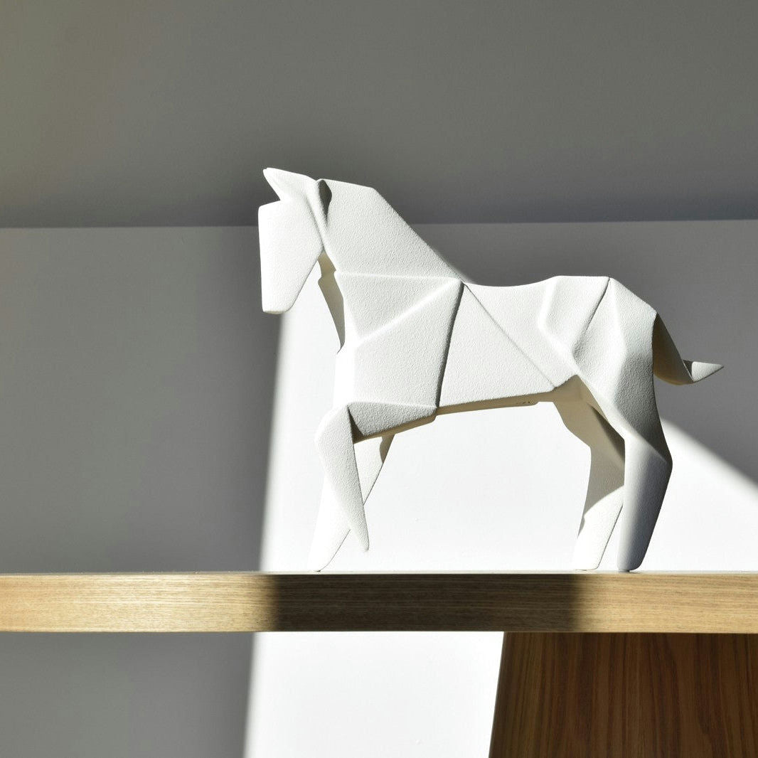 Una figura decorativa en cerámica Blanco mate Caballo Origami descansa elegantemente sobre un estante de madera, con la luz del sol proyectando una sombra en la pared detrás de ella.