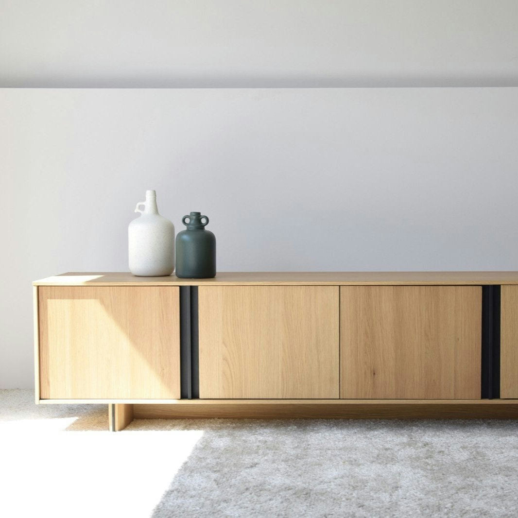 El mueble TV en DM y roble Clivo con detalles en metal negro se destaca contra una pared blanca sobre una alfombra gris claro, rematada con dos jarrones de cerámica, uno blanco y otro verde oscuro, para un elegante diseño contemporáneo.