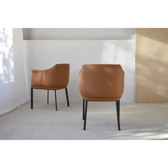 Set 2 Sillas Comedor Brazos Berlín: Dos modernas sillas de cuero marrón con patas negras, situadas sobre una alfombra clara en una habitación minimalista con paredes blancas y luz solar parcial, proyectando una elegancia contemporánea.