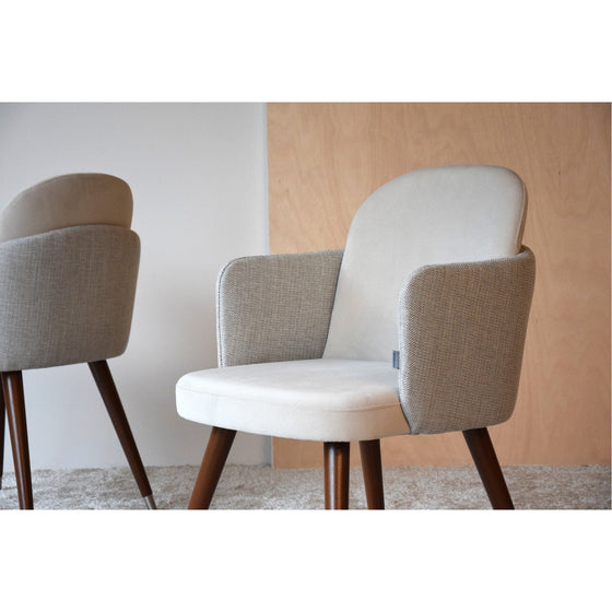 Dos sillas de comedor tapizadas con asiento beige y patas de madera del Pack 2 Sillas de Comedor Tapizadas con Reposabrazos Comodisigno se colocan sobre una alfombra en color claro y sobre un fondo de pared blanco y madera, mostrando su sofisticado diseño.