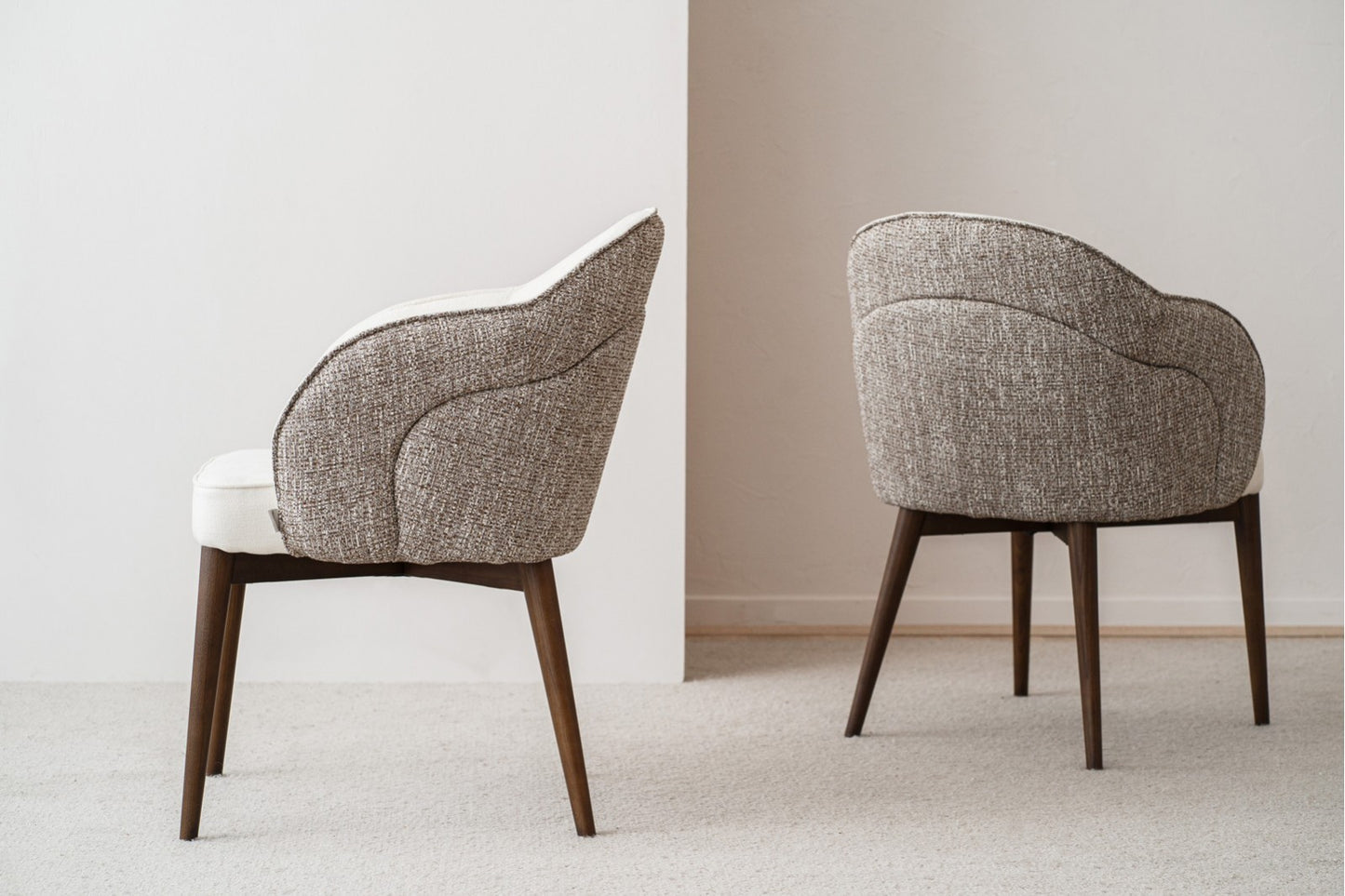 Un set de 2 sillas de comedor ROMA con brazos, tapizadas en tonos tostado y marfil y patas de madera nogal (55×60×83 cm), se ubica sobre una alfombra clara, una orientada a la izquierda y otra en ángulo, contra una pared neutra.