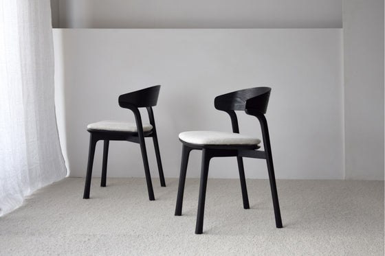 El Pack 2 Sillas de Comedor en Madera Emma, con modernos marcos negros y cojines claros, está colocado sobre un piso alfombrado beige contra una pared neutra, mostrando un diseño contemporáneo.