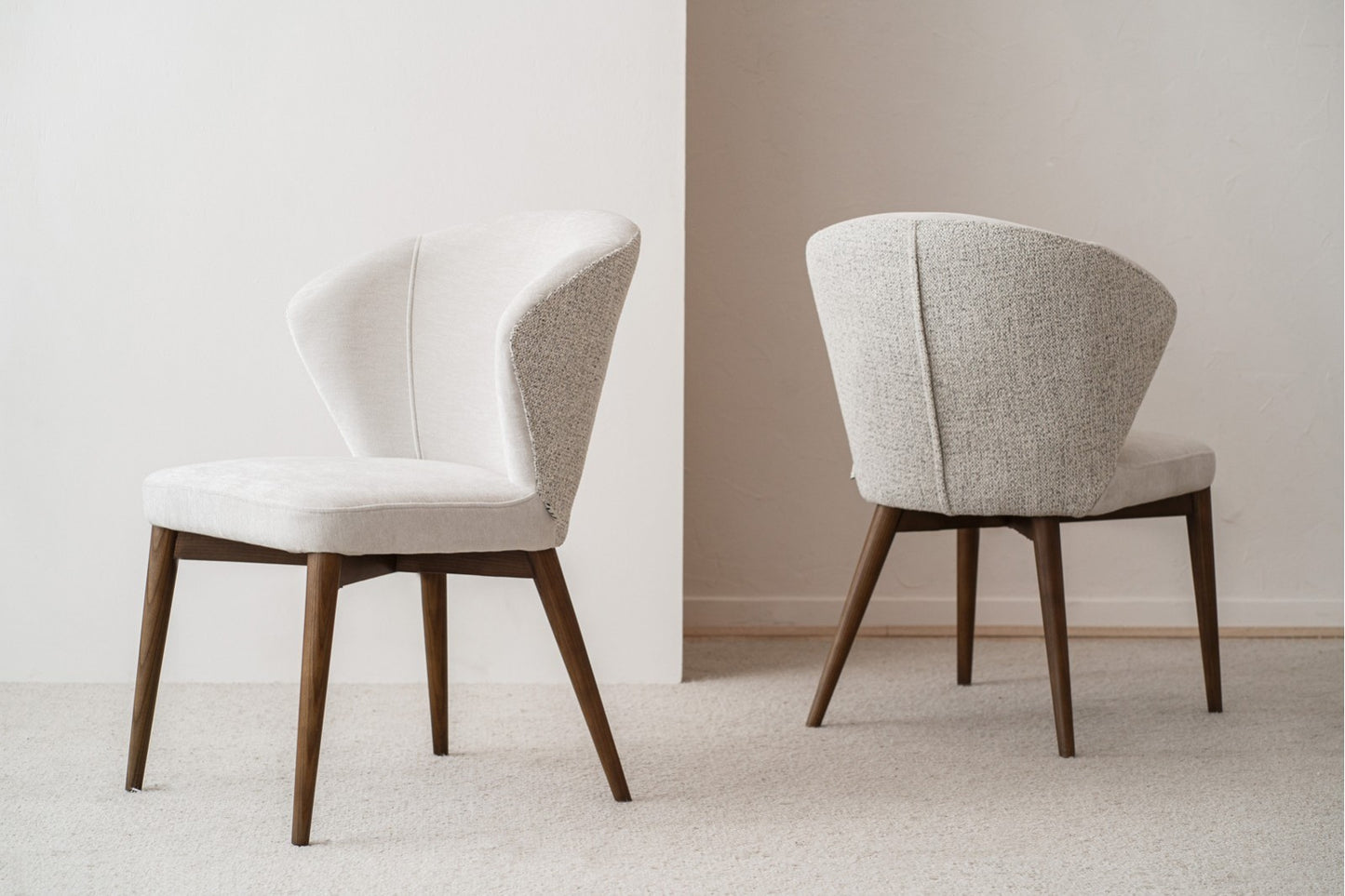 El Set de 2 sillas de comedor tapizadas en EVA beige y gris, con patas de madera nogal, está dispuesto sobre una alfombra clara: una silla de frente y la otra mostrando su respaldo.