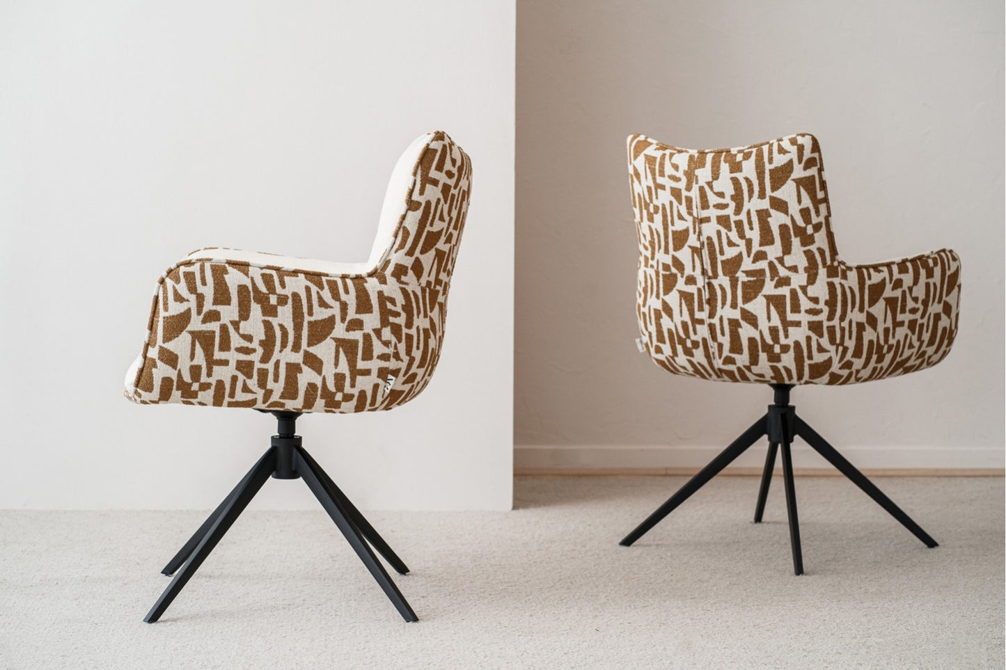 Dos sillas de comedor contemporáneas SAEL con reposabrazos, tapizadas en beige y ocre, presentan patas de metal negro. Las sillas giratorias (61×63×86 cm) están colocadas sobre una alfombra clara cerca de una pared blanca.
