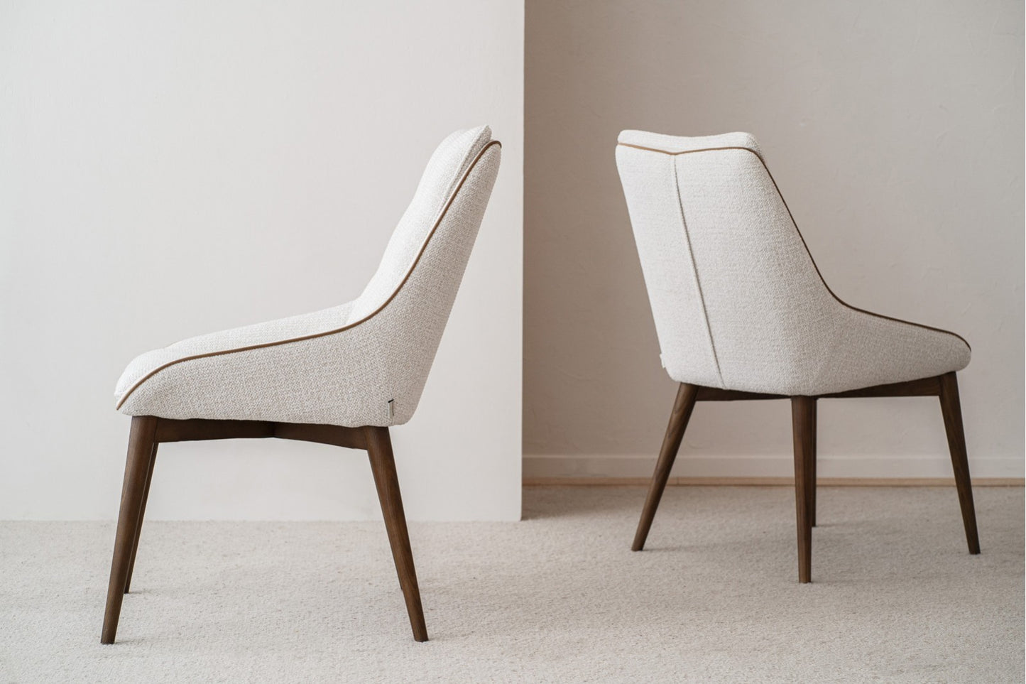 El set de 2 sillas de comedor MÍA, tapizadas en marfil (51×67×88 cm) y con patas de madera nogal, destaca sobre alfombra clara y fondo neutro. Ideales para comedor o para añadir un toque mid century a tu espacio.