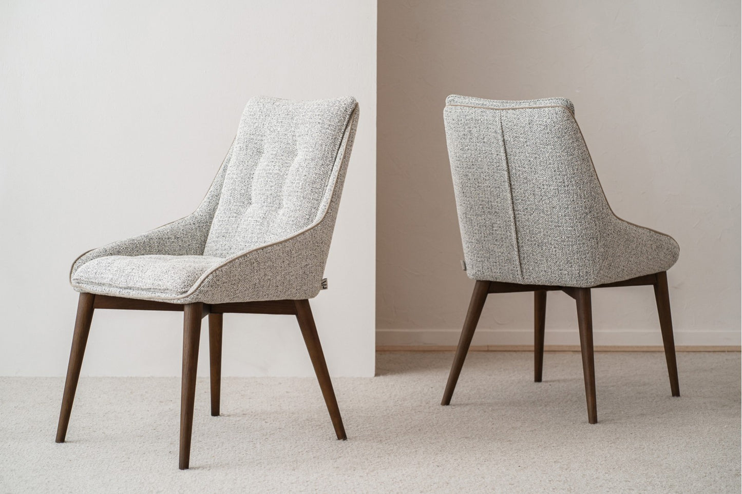 El set de 2 sillas de comedor tapizadas MÍA en tonos grises (51×67×88 cm) con estructura de madera nogal estilo mid century está colocado en una sala minimalista con paredes neutras y alfombra.