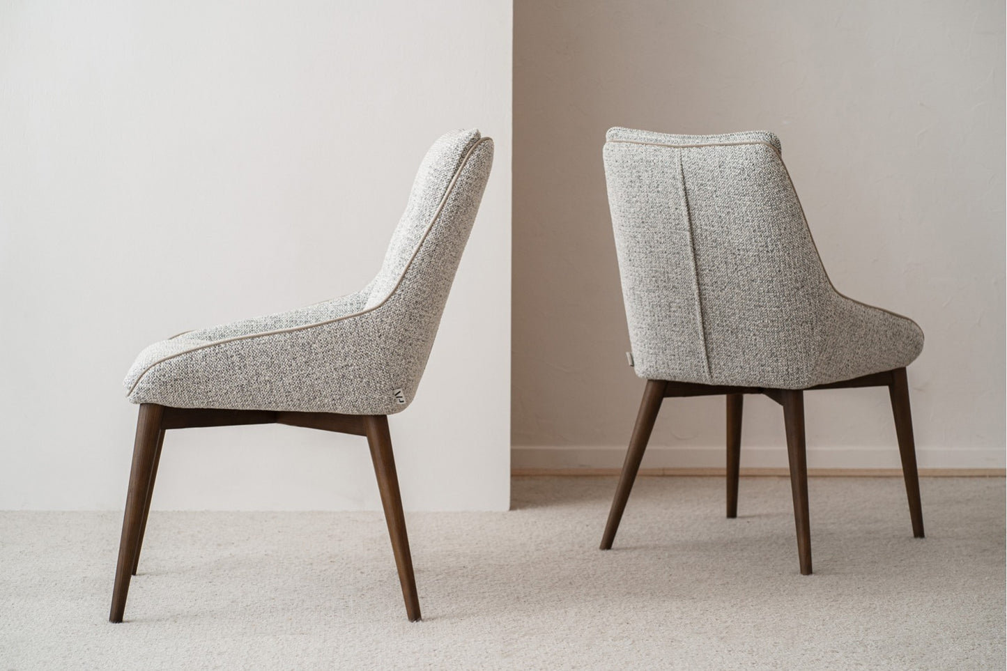 El set de 2 sillas de comedor MÍA, tapizadas en tonos grises y con patas de madera nogal (51×67×88 CM), está dispuesto espalda con espalda en una habitación minimalista de tonos grises y moqueta neutra.