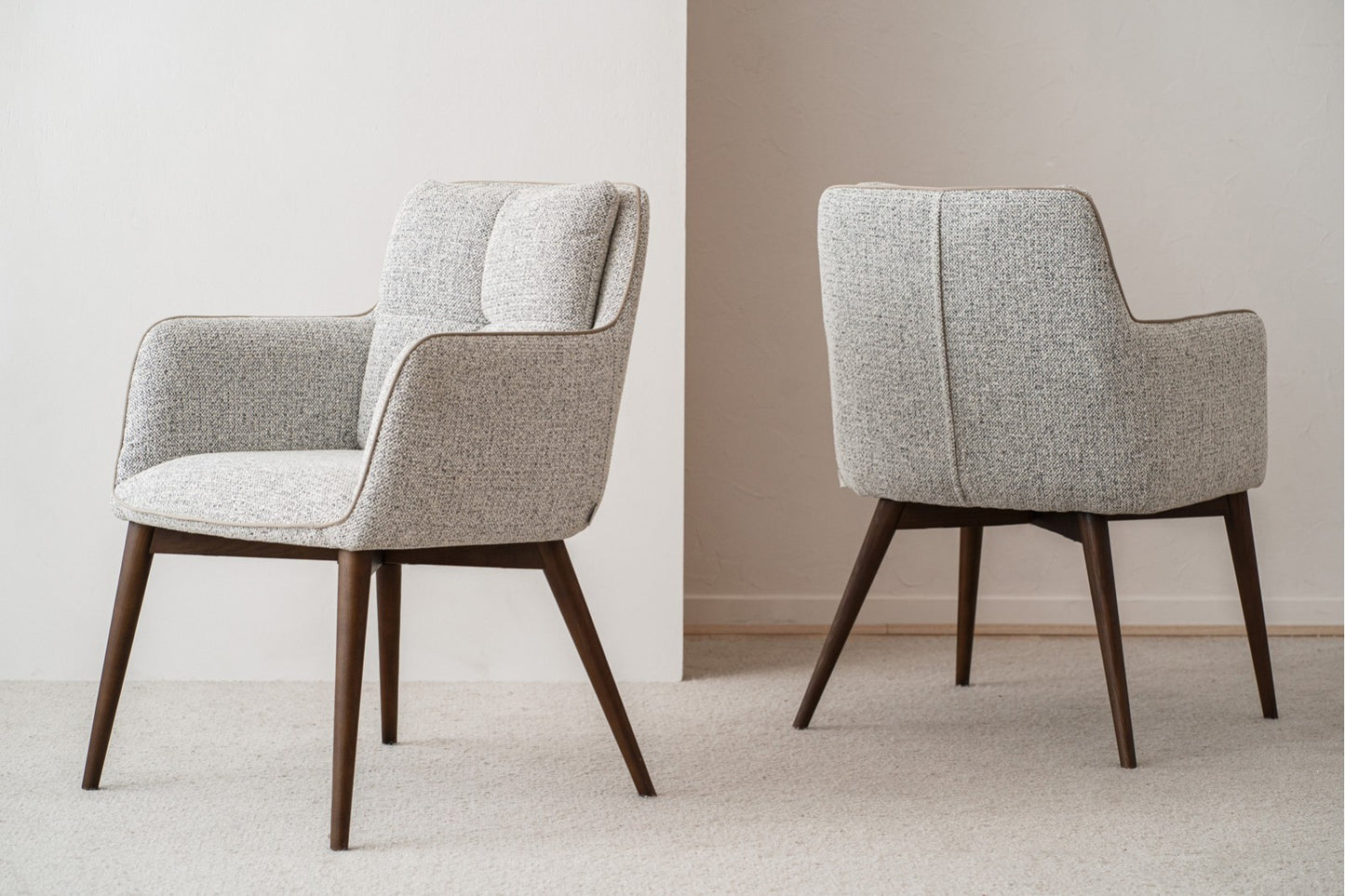 El set de 2 sillas de comedor OLIVER, tapizadas en tono gris con brazos y madera nogal (61×67×86 cm), está ubicado sobre una alfombra clara junto a una pared blanca.
