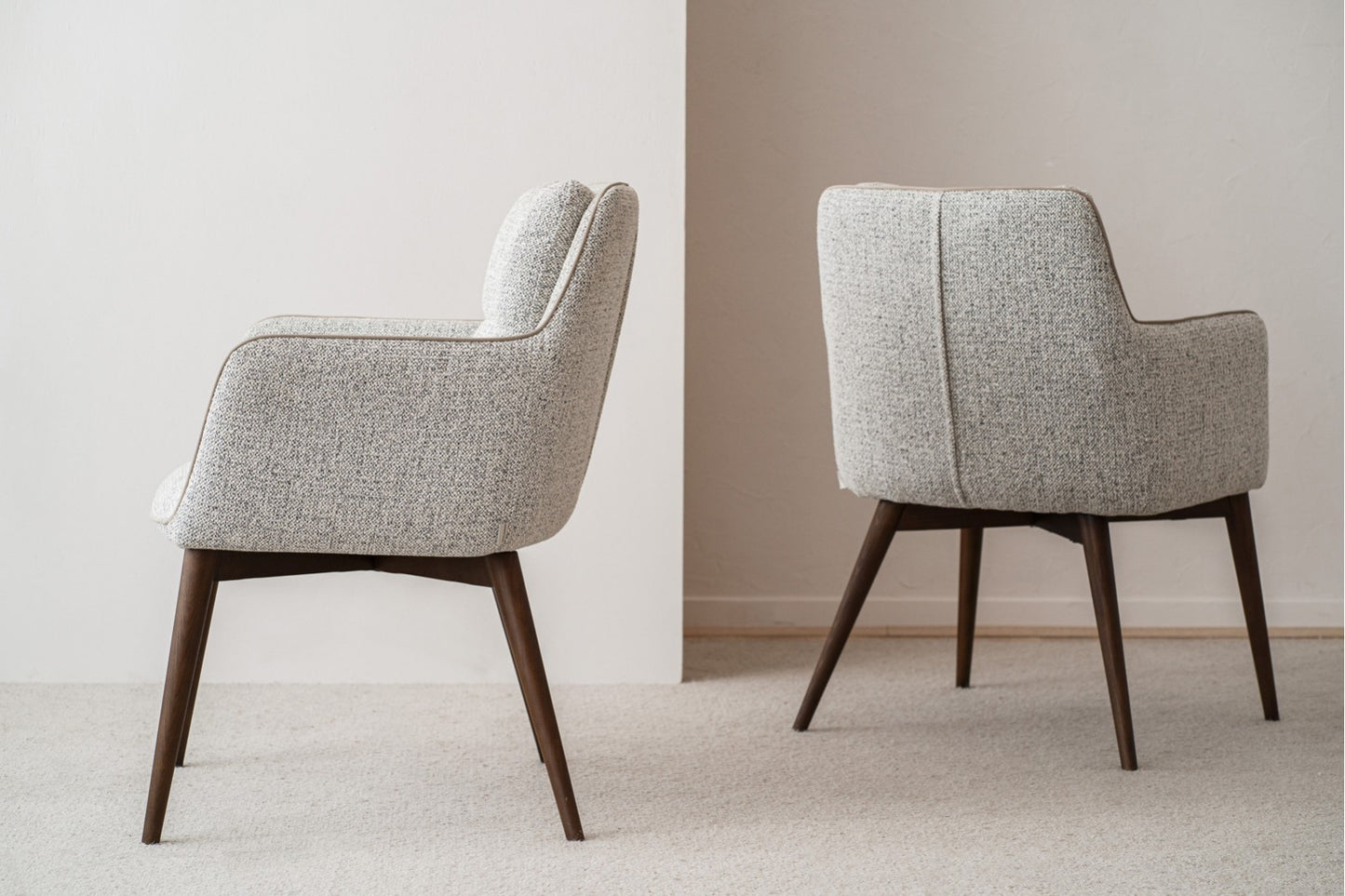 El set de 2 sillas de comedor OLIVER, tapizadas en gris y con patas de madera nogal (61×67×86 cm), está colocado sobre una alfombra frente a una pared blanca, ligeramente separadas entre sí.