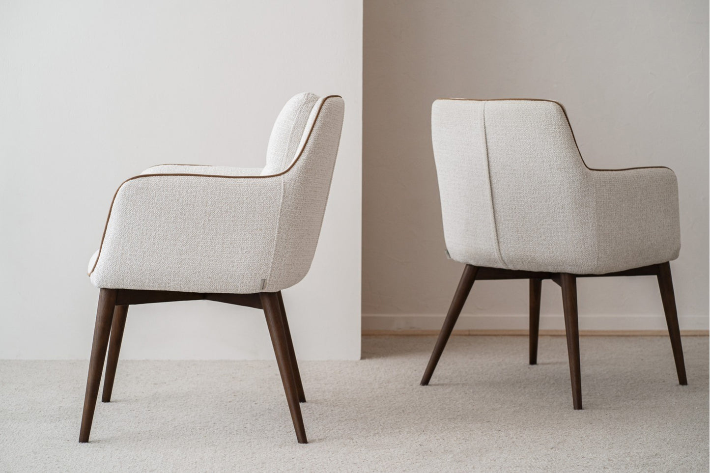 El set de 2 sillas de comedor OLIVER con brazos, tapizadas en tono marfil y patas de madera nogal (61×67×86 cm), se ubica sobre alfombra clara frente a una pared neutra, orientadas en direcciones opuestas.