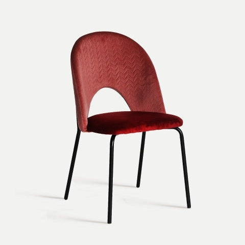 La silla de comedor en terciopelo y hierro Sarkad, una silla de tela roja que refleja un diseño vintage moderno, presenta un respaldo curvo y elegantes patas de metal negro sobre un fondo blanco liso.