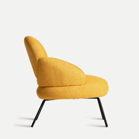 El sillón de chenilla mostaza Dokkum irradia sofisticación moderna con su tapizado amarillo, respaldo curvo y apoyabrazos elegantemente colocados sobre elegantes patas de metal negro sobre un fondo liso.