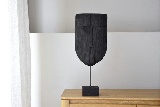 La figura decorativa en madera de mango Máscara de la Colección Máscara reposa elegantemente sobre una superficie de madera. Su soporte acentúa su forma oscura y alargada contra una pared sencilla y de color claro con una cortina parcialmente visible a la izquierda, que combina el arte tradicional con el encanto moderno.