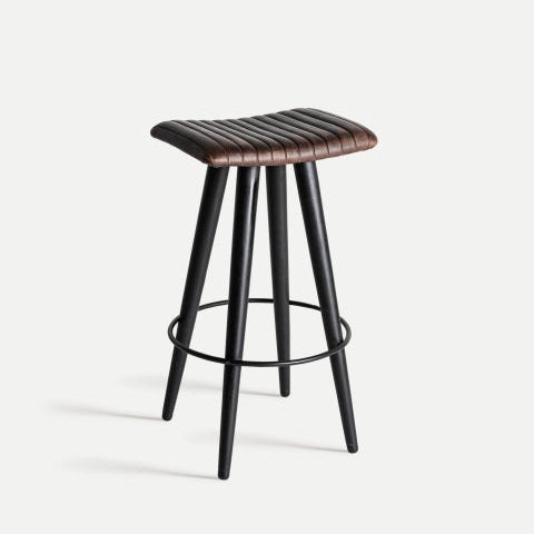 El Taburete alto 74 cm en piel y metal Befaux combina un asiento acanalado marrón con cuatro patas negras y un reposapiés circular, incorporando un aire de diseño industrial vintage.