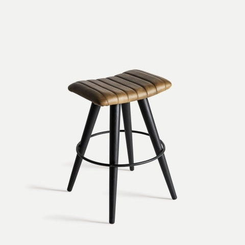El Taburete en piel y metal Befaux presenta un asiento acanalado y acolchado de color marrón con patas negras y un reposapiés circular, que incorpora un diseño contemporáneo industrial sobre un fondo blanco liso.