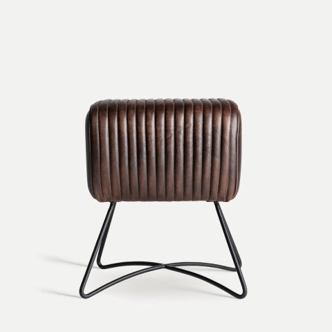 El Taburete bajo de piel y base metálica Gladys, con su asiento cilíndrico de cuero marrón y textura acanalada, encarna el diseño industrial moderno. Sus elegantes patas de metal negro realzan una cocina abierta con estilo y funcionalidad sobre un fondo liso.