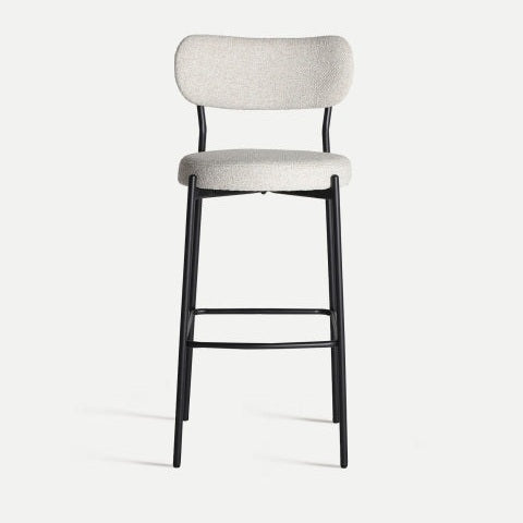 El Taburete alto Segonzac es un taburete alto con un asiento tapizado en gris claro y un respaldo de apoyo sobre un elegante marco de metal negro, sobre un fondo blanco liso.