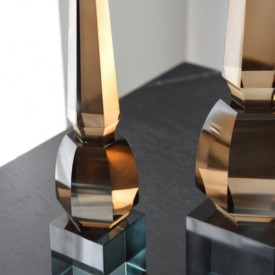 Las "Figuras decorativas en Cristal Pulido Obelisco", dos objetos altos de vidrio marrón con formas geométricas y superficies reflectantes elaborados con artesanía meticulosa, se colocan sobre una mesa oscura y brillante.