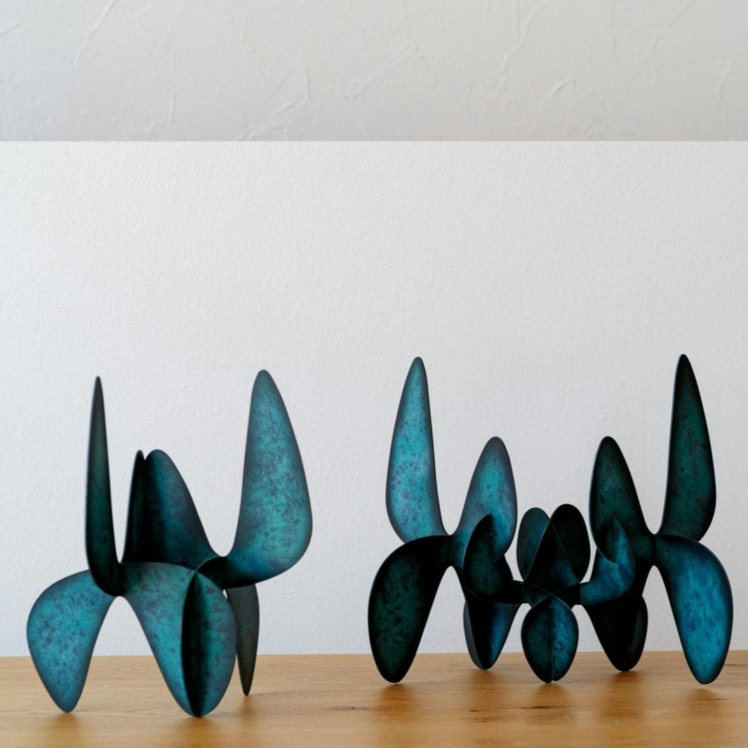Dos esculturas abstractas y curvas de metal en color verde azulado descansan sobre una superficie de madera sobre un fondo blanco, resaltando la elegancia de la colección Figuras Decorativas en Metal Orgánico Lámina.