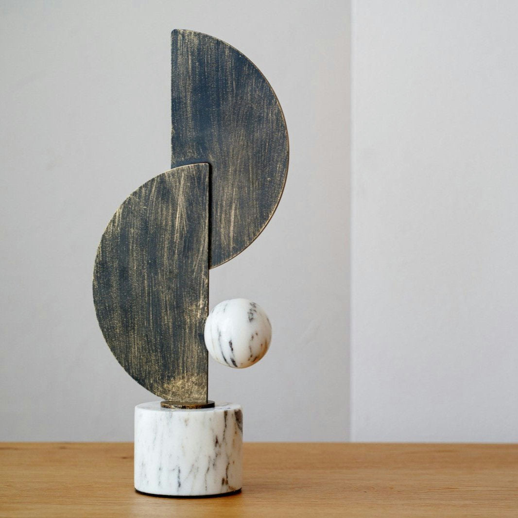 Las Figuras decorativas en mármol y metal Arco N1 es una escultura abstracta con semicírculos verticales en negro y gris, una esfera de mármol y una base cilíndrica sobre madera. Esta pieza única es ideal para la decoración de interiores con sus elegantes detalles en mármol blanco.