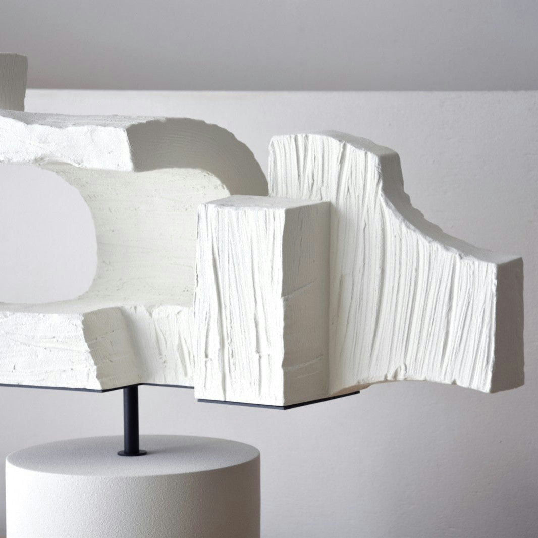 La Figura Decorativa en Cerámica Blanca Hormión es una escultura abstracta blanca con una superficie texturizada, que presenta formas geométricas y curvas, montada sobre un pedestal cilíndrico sobre un fondo liso, que encarna un diseño versátil.
