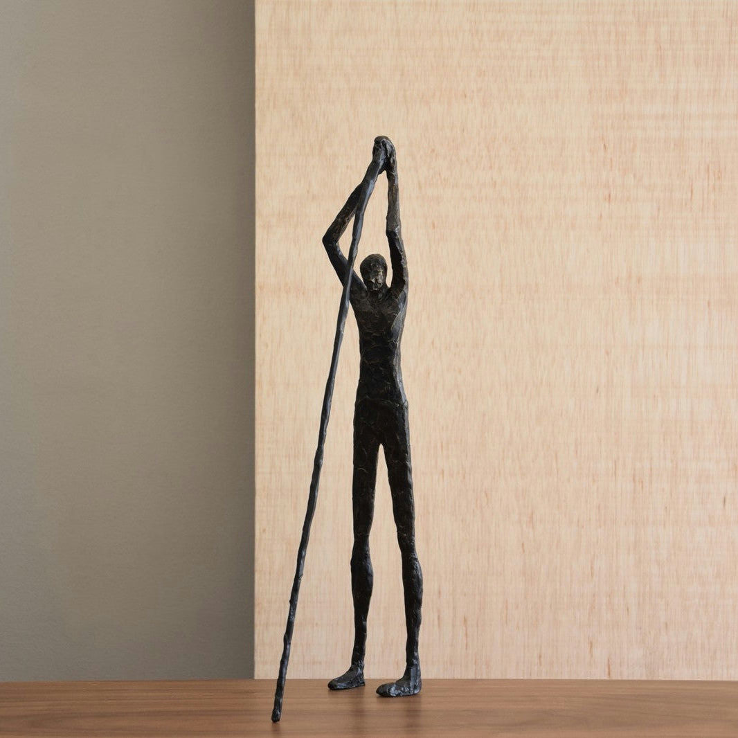 Una figura decorativa de metal en bronce envejecido Hombre N1, esbelta y alargada, representa a una persona sosteniendo un palo largo, de pie sobre una superficie de madera con un fondo neutro, que encarna el diseño minimalista y ejemplifica el arte de calidad.