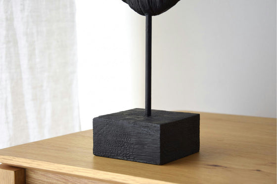 Una escultura de madera de mango negro, llamada Figura decorativa en madera de mango Máscara, presenta una varilla vertical sobre una base rectangular y se exhibe elegantemente como parte de la prestigiosa Colección Máscara.