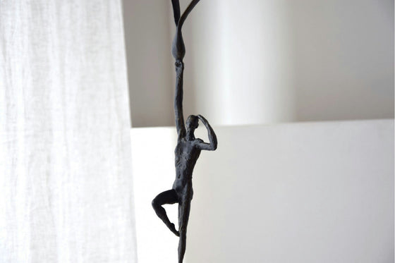 Una esbelta escultura abstracta en bronce y mármol, que representa una figura humana trepando o estirándose sobre un fondo claro con cortinas blancas transparentes, encarna la elegancia de Figura Decorativa en Bronce y Mármol Horizonte.