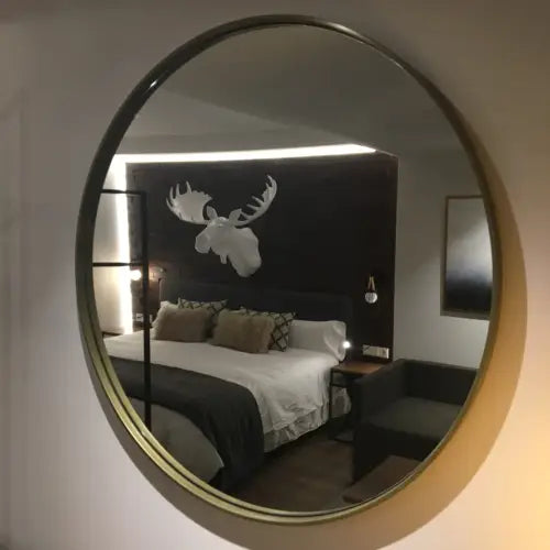 El Espejo Circular Eye refleja una imagen de un dormitorio con cabeza de alce decorativa sobre la cama, presentando un diseño minimalista.